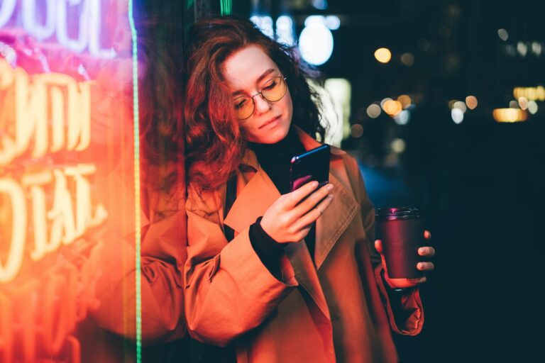 Successful woman using smartphone outdoors while standing near skyscraper at night.